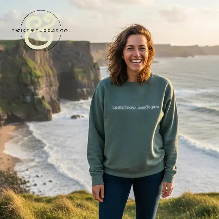Smiling woman in custom embroidered sweatshirt reading 'Tomorrow needs you' by Twist & Thread Co. near cliffs and ocean.