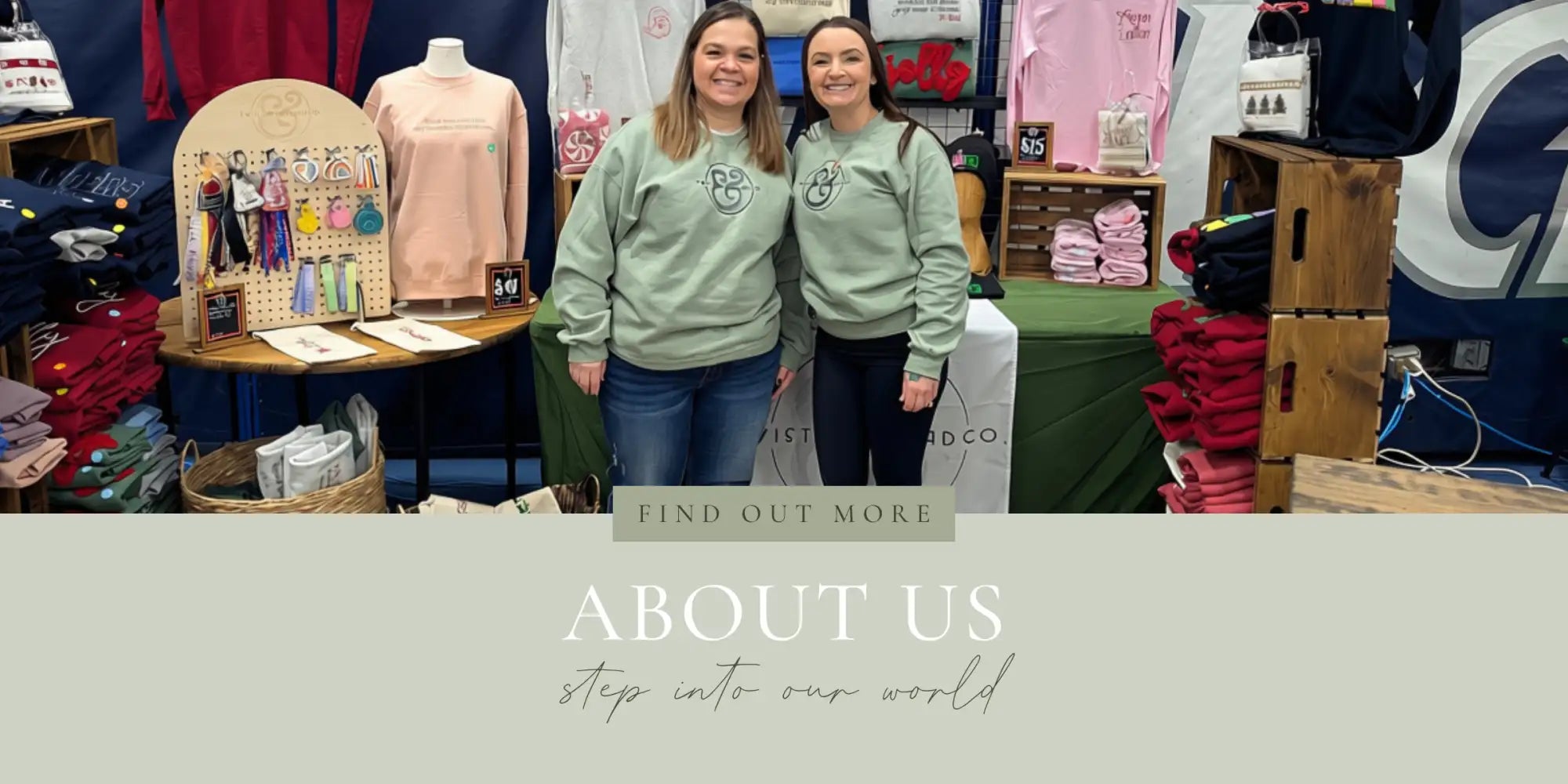 Two women in matching light green sweatshirts with a small embroidered logo on the chest stand together at what appears to be a boutique or pop-up shop.