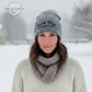 Woman wearing grey knit winter hat with embroidered text, scarf, and sweater in snowy outdoor setting