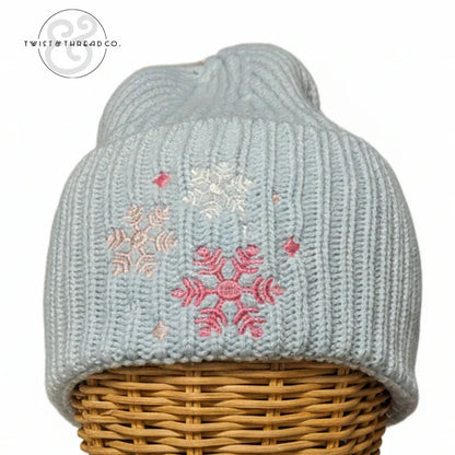 Light blue knit winter hat with pink and white snowflake embroidery on a wicker stand